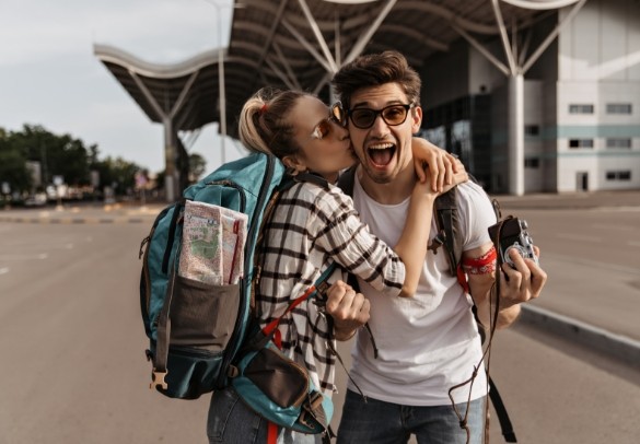 Pareja de viajeros felices frente a un aeropuerto; la mujer besa al hombre en la mejilla mientras ambos sonríen. Llevan mochilas, ropa casual y una cámara, listos para una nueva aventura.